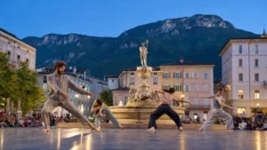 นักเต้นร่วมสมัยแสดงรอบน้ำพุ Piazza Rosmini เมืองโรเวเรโต โดยมีภูเขาเตรนติโนเป็นฉากหลัง